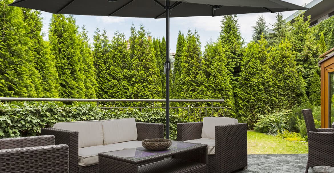 Gartenmöbel mit Sonnenschirm auf Terrasse, gepflegter Garten mit hohen Hecken, gemütliche Sitzgruppe aus Rattan, perfekte Gartengestaltung für Garten- und Landschaftsservice.